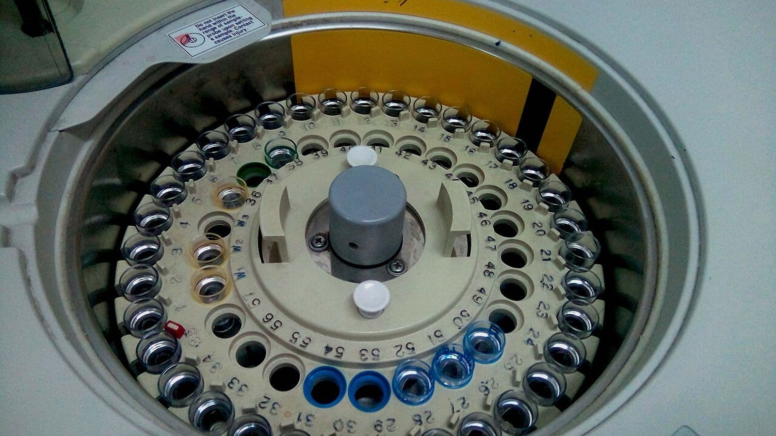 Carrossel circular numerado com dezenas de copinhos de amostra dentro de um analisador automático de laboratório