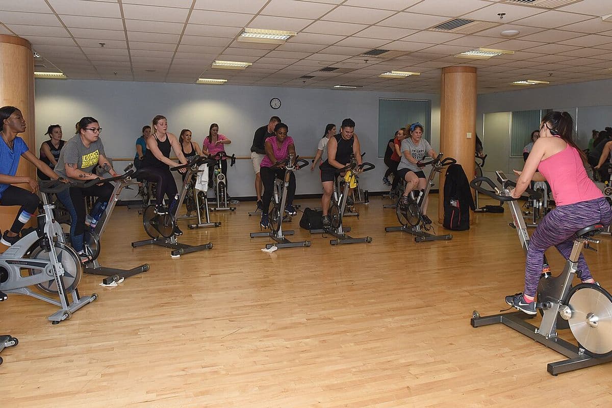Turma em aula de ciclismo indoor em sala com piso de madeira, com a instrutora de costas à direita