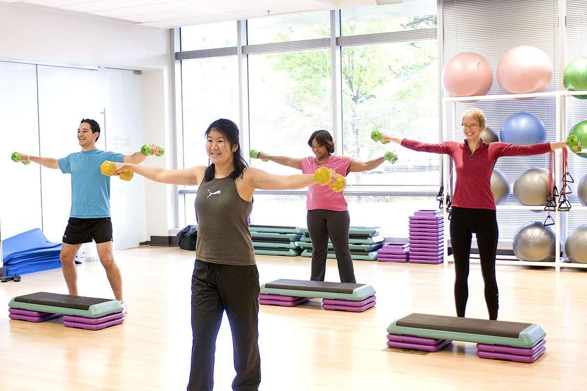 Quatro alunos de pé em uma sala de ginástica, com os braços abertos na lateral do corpo segurando halteres leves
