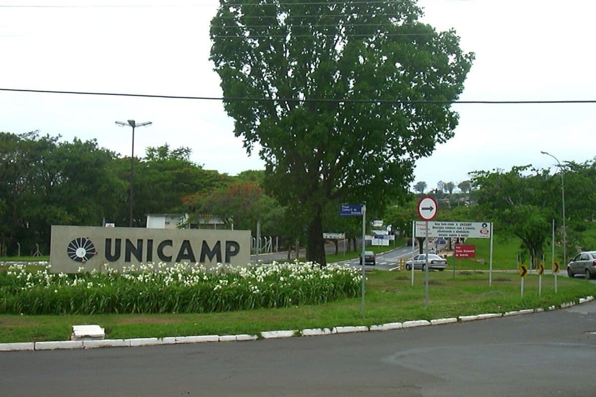 Letreiro com o nome Unicamp em um canteiro de flores na entrada do campus, com árvores e uma rua ao fundo