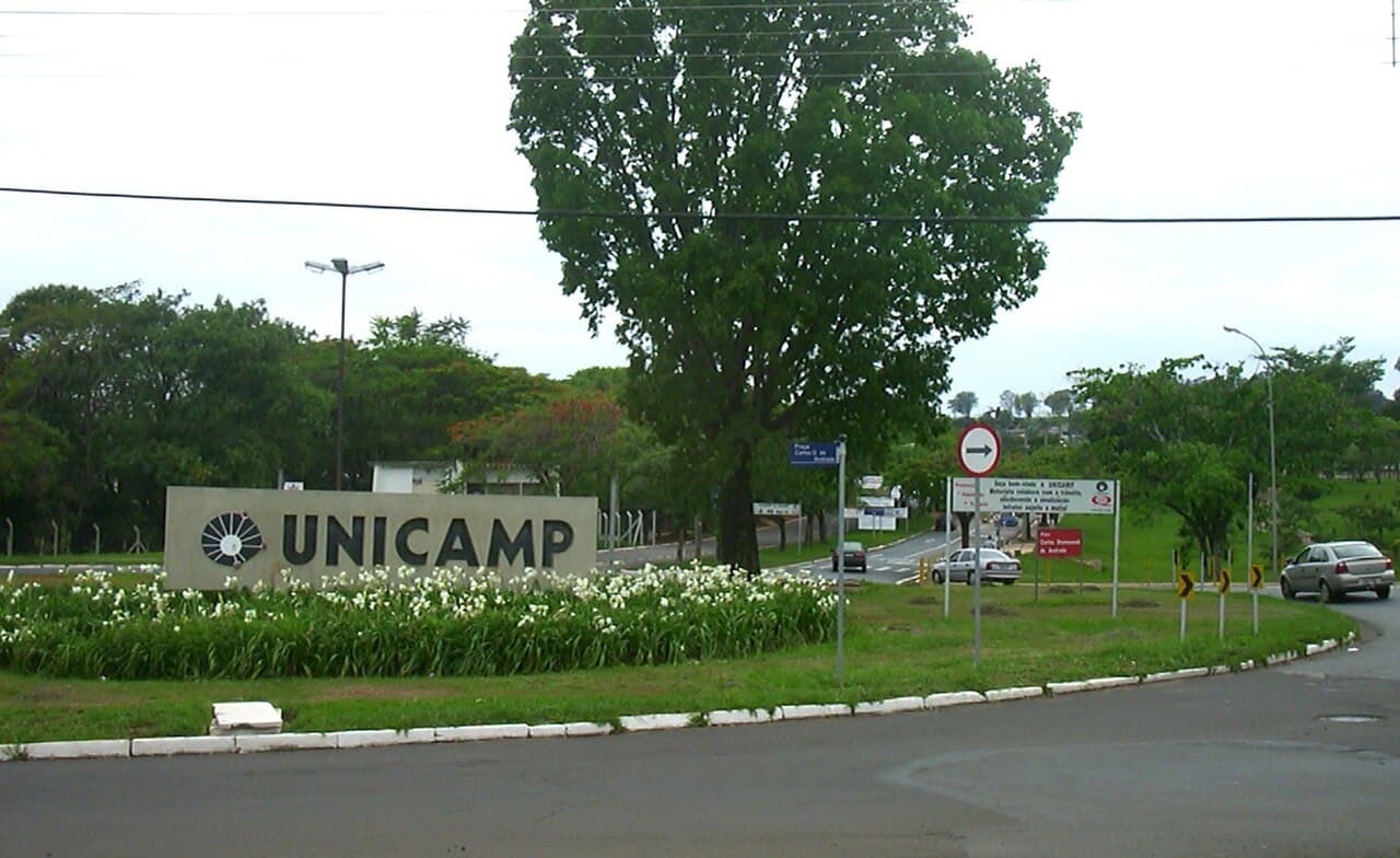 Letreiro com o nome Unicamp em um canteiro de flores na entrada do campus, com árvores e uma rua ao fundo