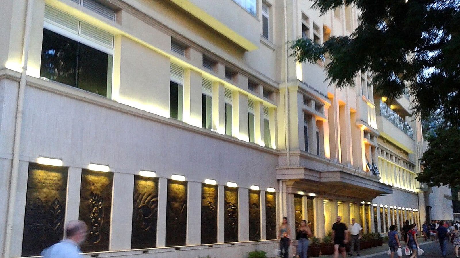Fachada iluminada de um prédio hospitalar de vários andares, com painéis em relevo no térreo e pedestres na calçada ao entardecer