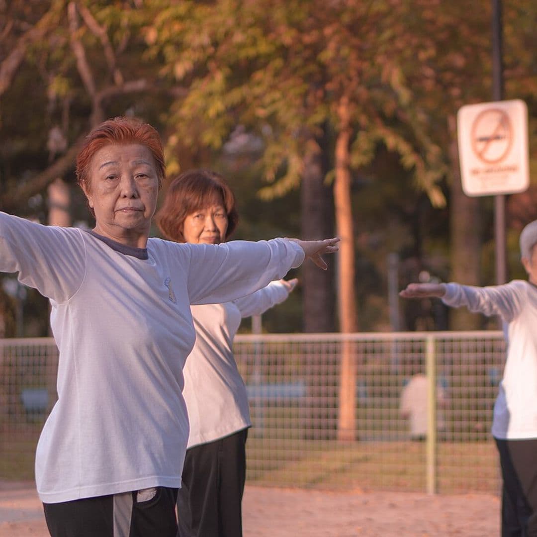 Três mulheres idosas de camiseta branca fazem tai chi com os braços abertos num parque