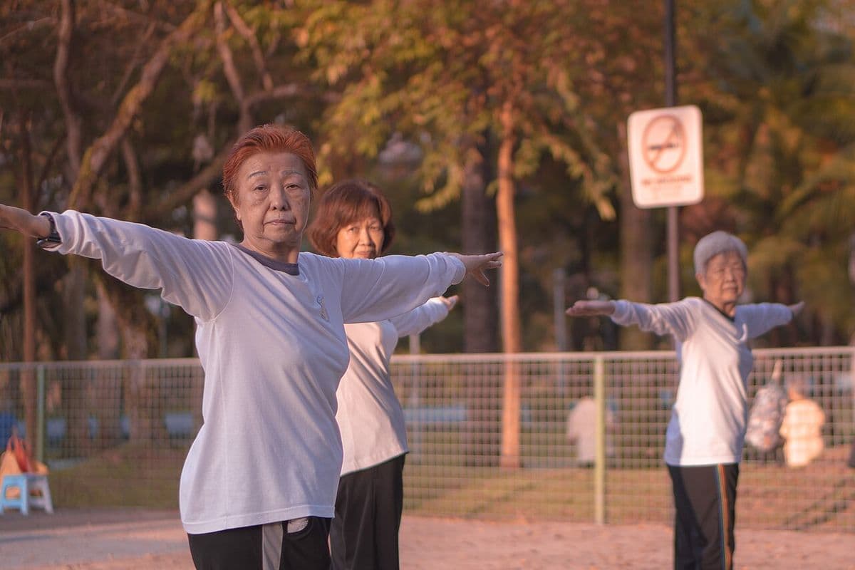 Três mulheres idosas de camiseta branca fazem tai chi com os braços abertos num parque