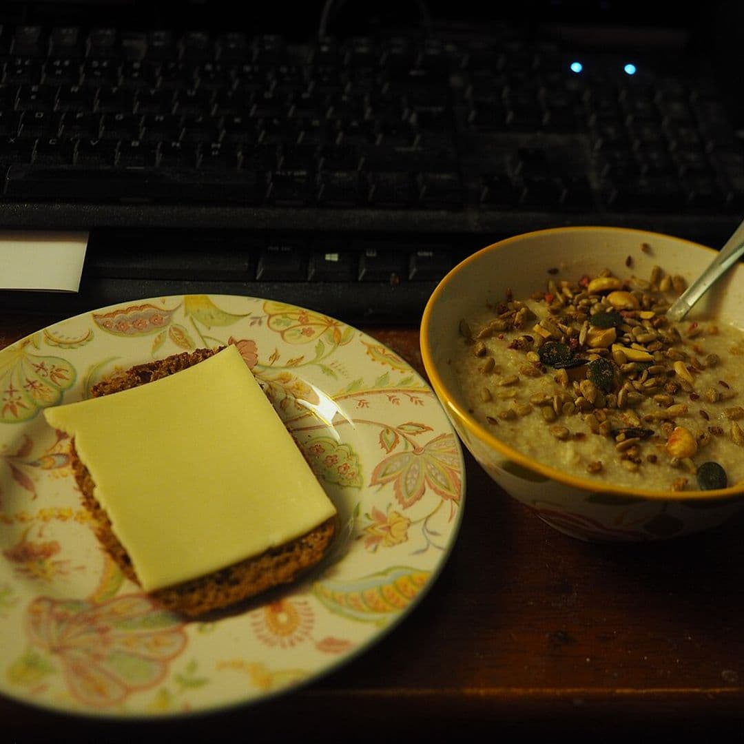 Prato com fatia de pão escuro e queijo ao lado de uma tigela de mingau de aveia com sementes, diante de um teclado de computador, em ambiente com pouca luz