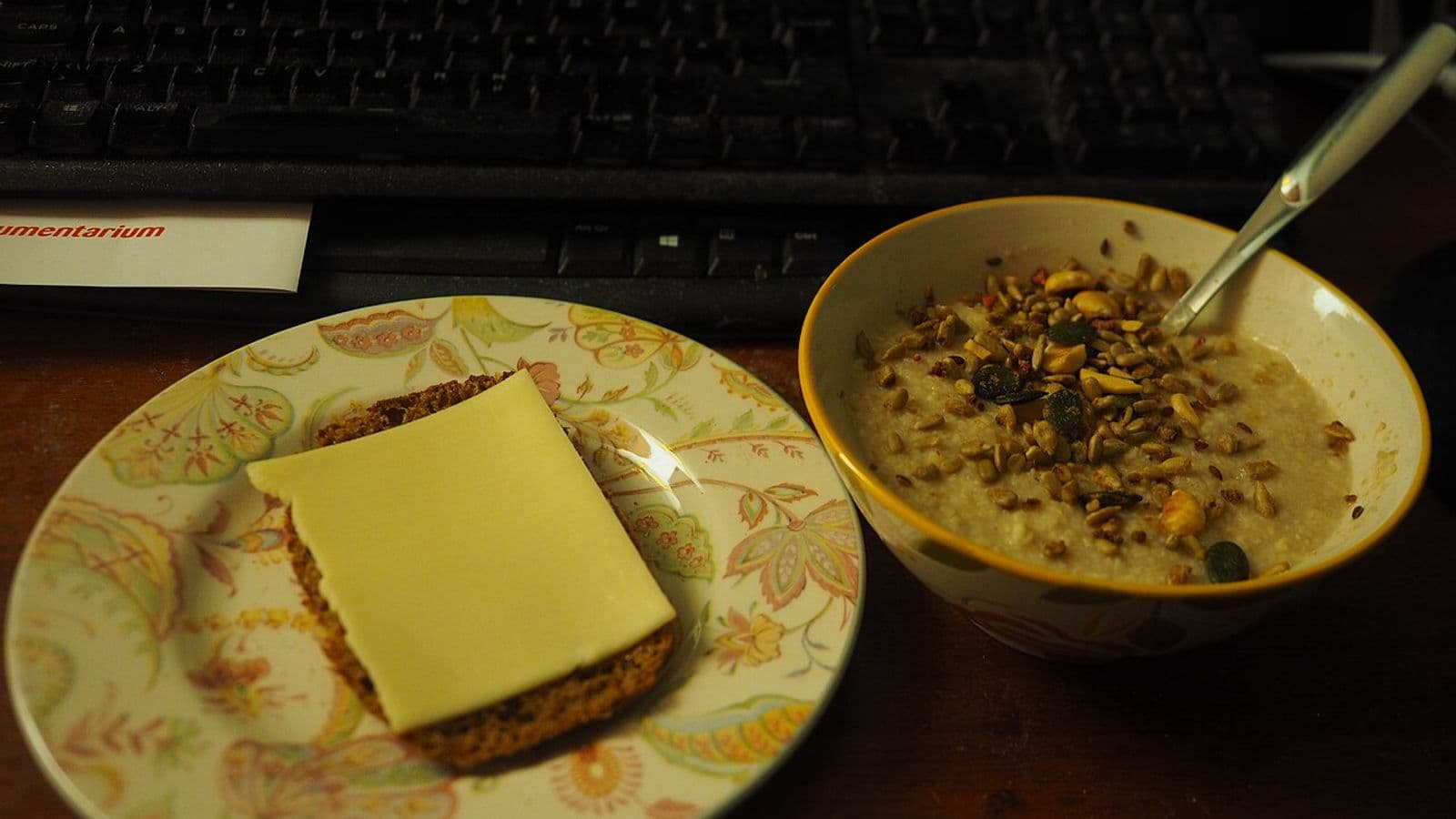 Prato com fatia de pão escuro e queijo ao lado de uma tigela de mingau de aveia com sementes, diante de um teclado de computador, em ambiente com pouca luz