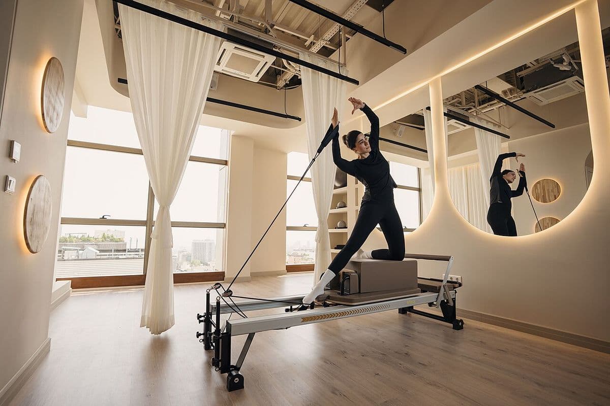 Mulher de roupa preta ajoelhada sobre a caixa de um reformer, segurando a alça, em um studio de pilates com janelas amplas e espelho