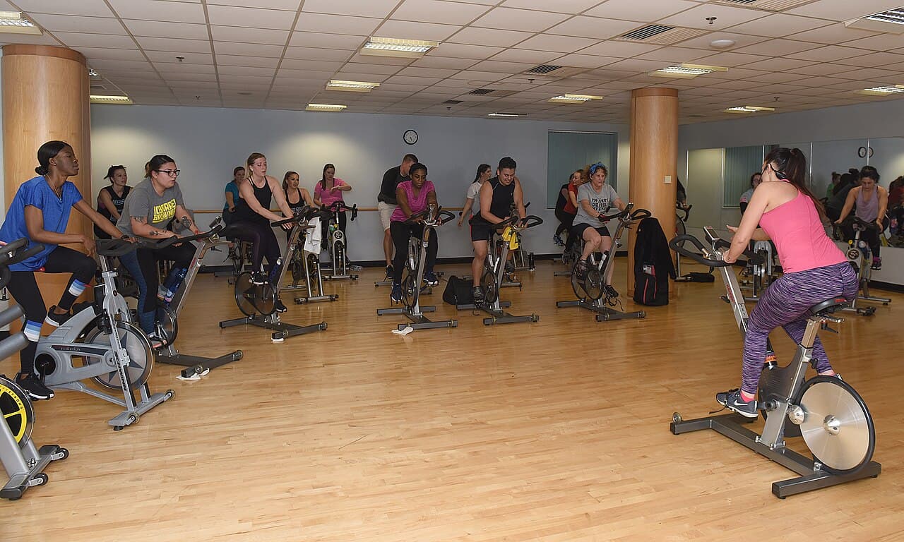 Turma em aula de ciclismo indoor em sala com piso de madeira, com a instrutora de costas à direita
