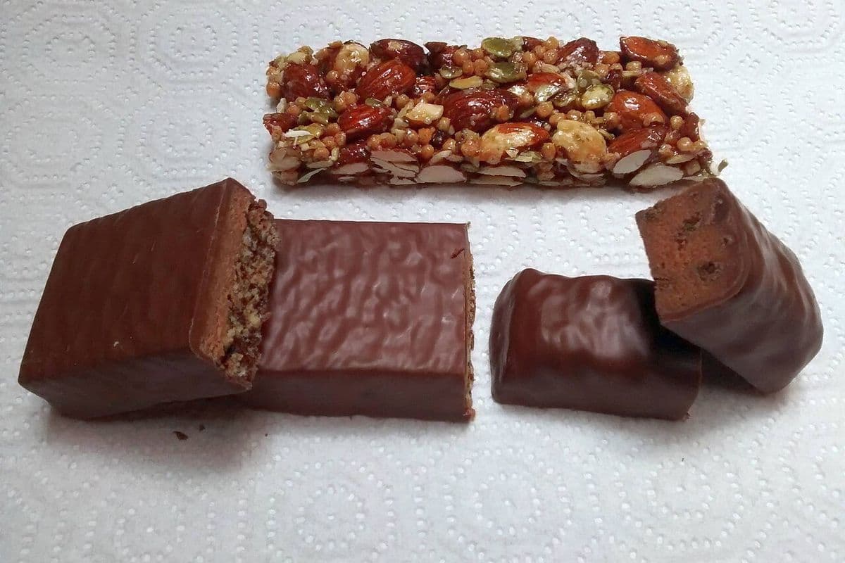 Três barras de proteína sem embalagem sobre papel-toalha: uma de castanhas e duas cobertas de chocolate, cortadas ao meio