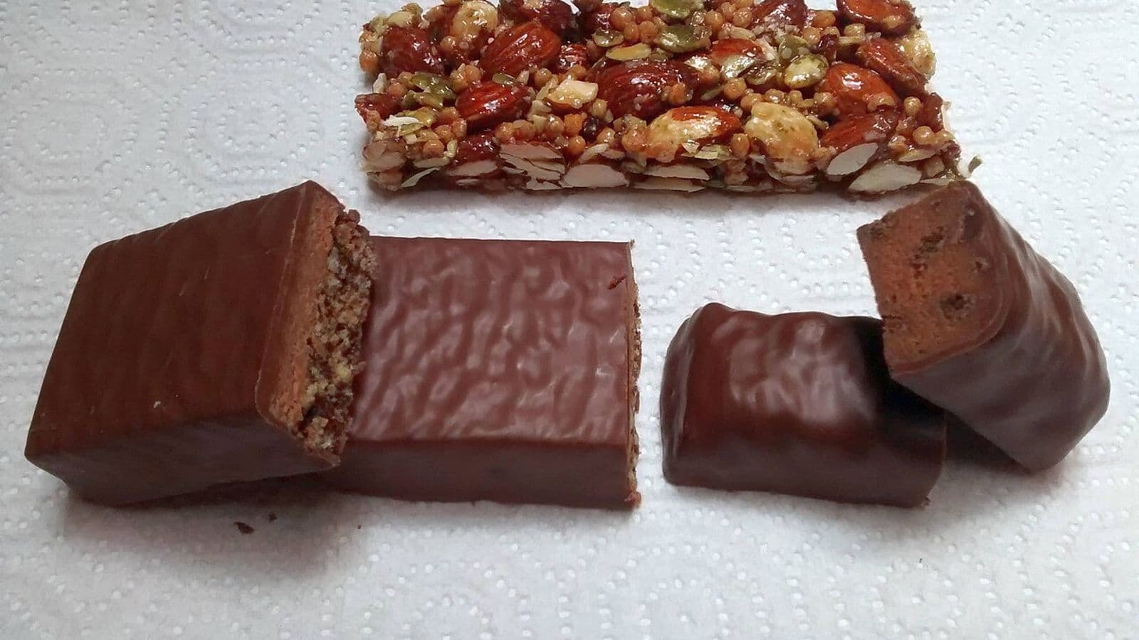 Três barras de proteína sem embalagem sobre papel-toalha: uma de castanhas e duas cobertas de chocolate, cortadas ao meio