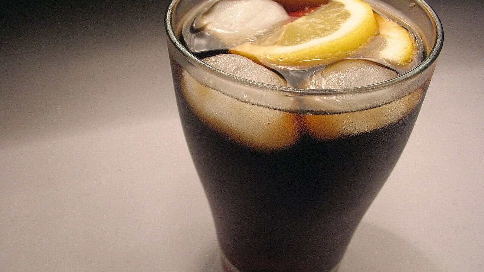 Copo de vidro com refrigerante de cola, cubos de gelo e duas fatias de limão sobre fundo cinza