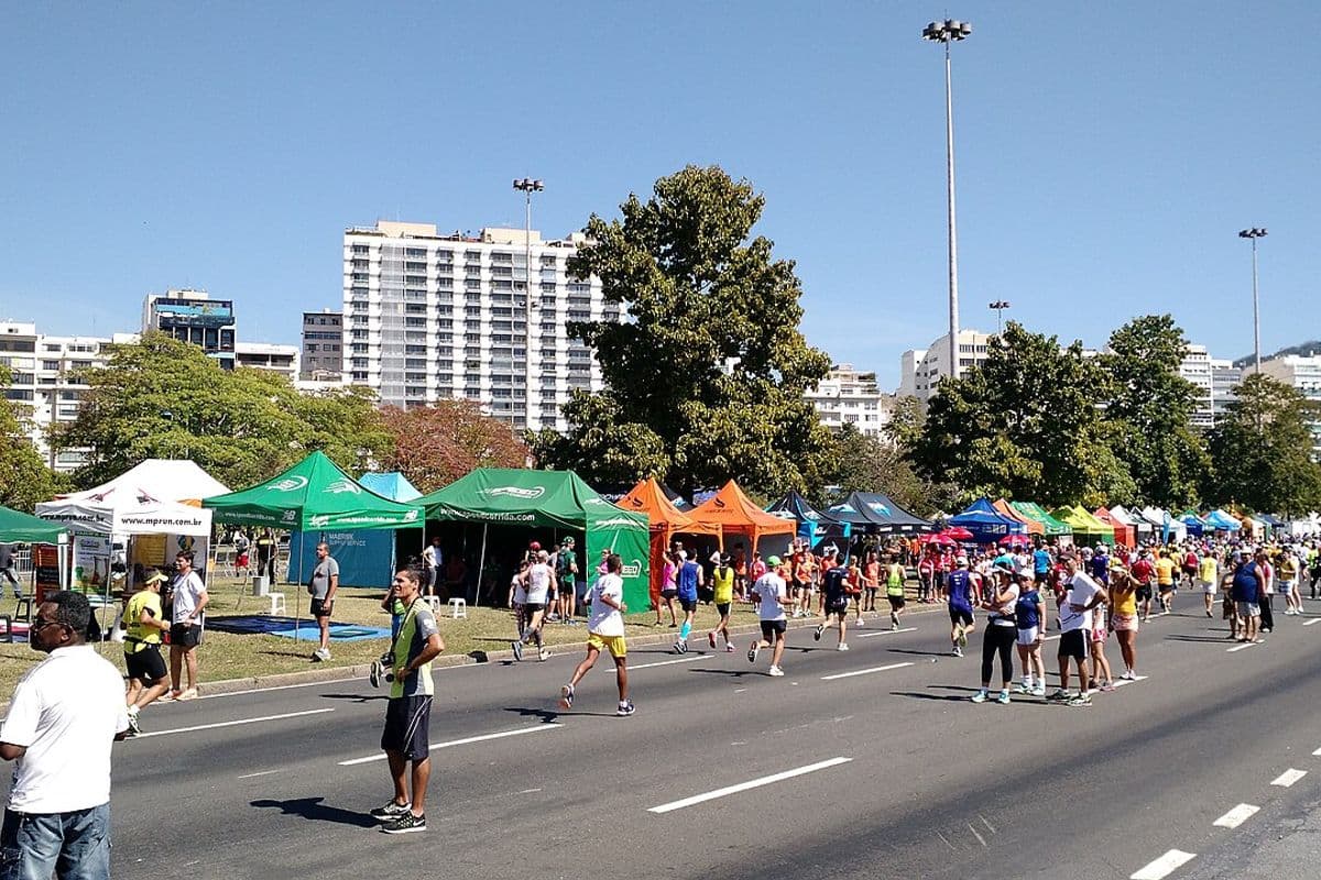 Corredores amadores numa avenida do Rio de Janeiro, com tendas coloridas de equipes de corrida e prédios ao fundo