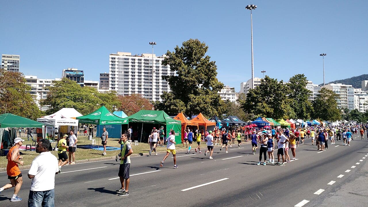 Corredores amadores numa avenida do Rio de Janeiro, com tendas coloridas de equipes de corrida e prédios ao fundo