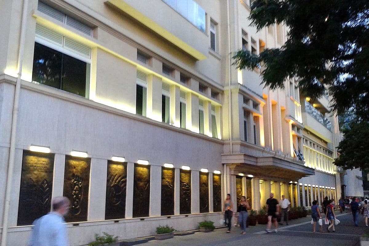 Fachada iluminada de um prédio hospitalar de vários andares, com painéis em relevo no térreo e pedestres na calçada ao entardecer