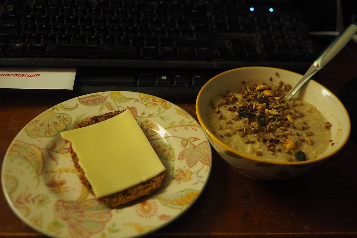Prato com fatia de pão escuro e queijo ao lado de uma tigela de mingau de aveia com sementes, diante de um teclado de computador, em ambiente com pouca luz