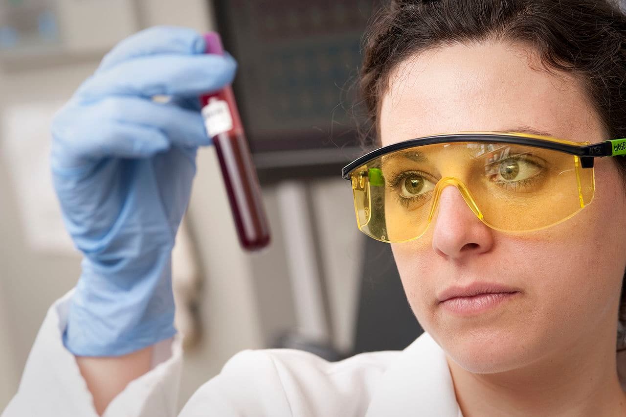 Pesquisadora de óculos de proteção e luva azul observa um tubo com amostra de sangue no laboratório