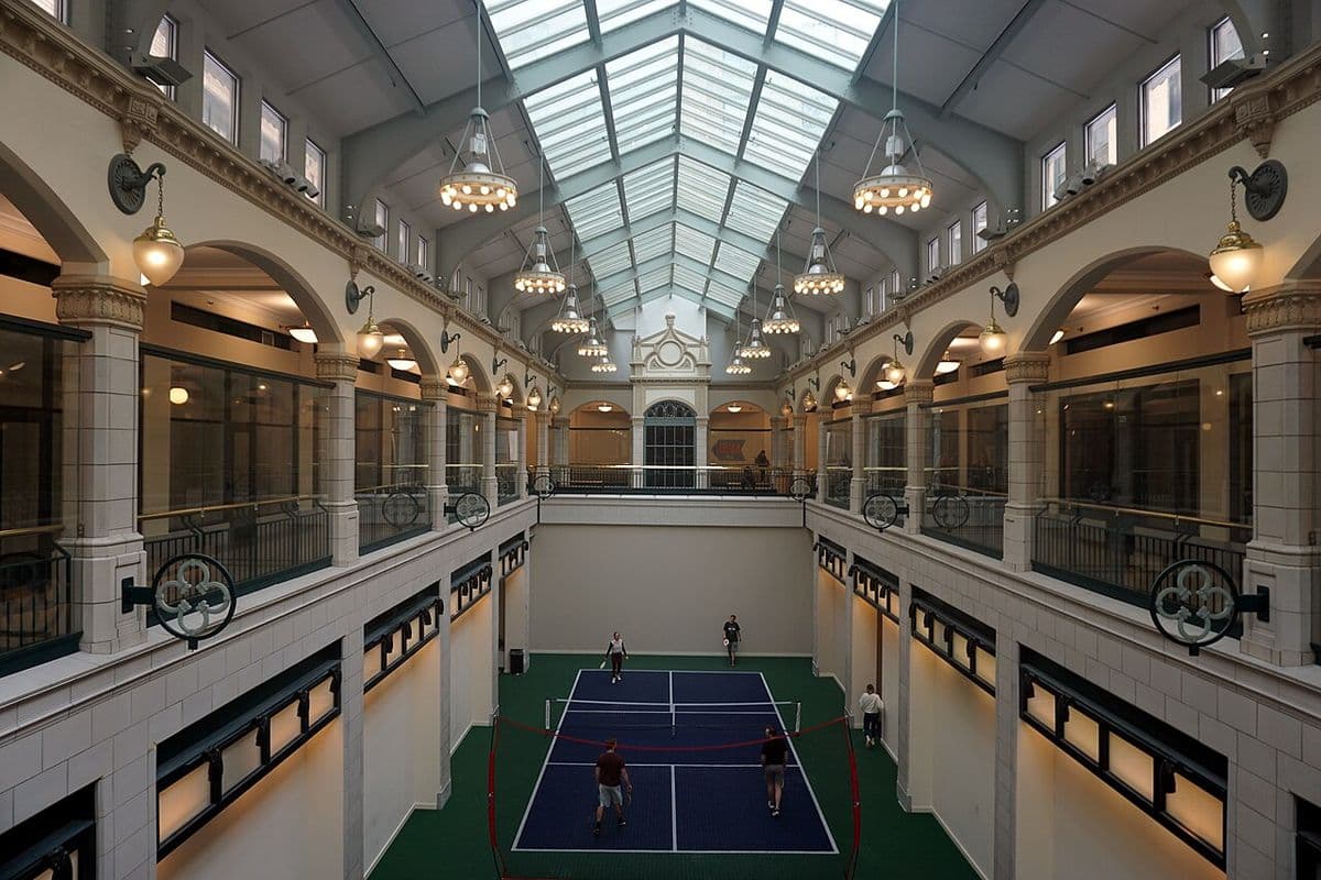 Quadra de pickleball coberta no átrio de um prédio histórico, com quatro jogadores e claraboia de vidro no teto