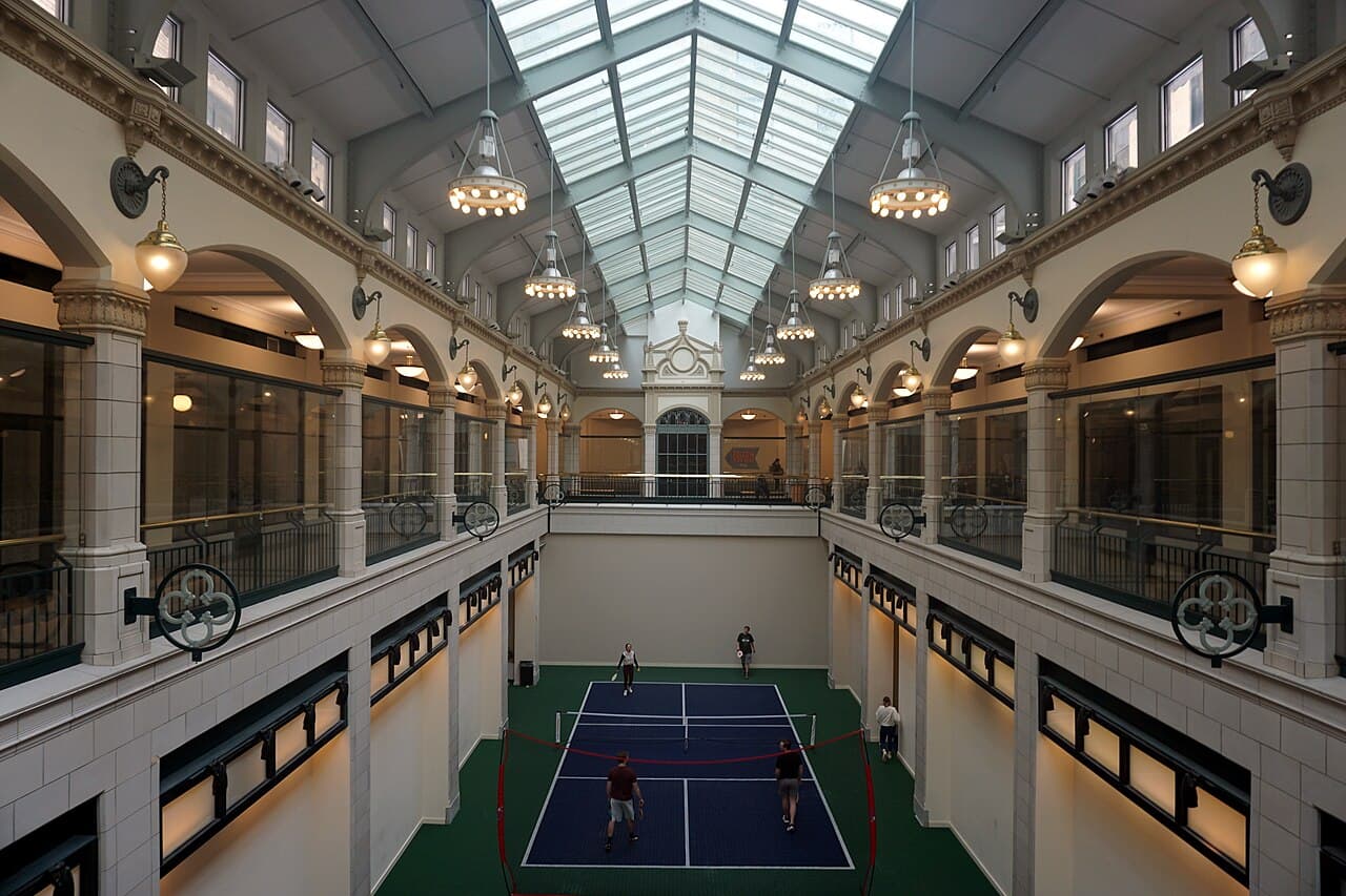 Quadra de pickleball coberta no átrio de um prédio histórico, com quatro jogadores e claraboia de vidro no teto