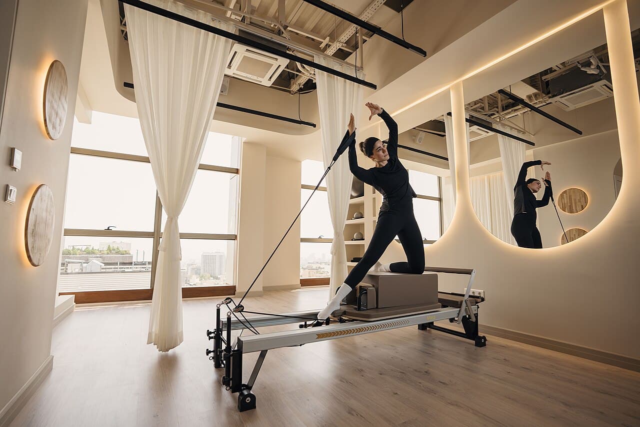 Mulher de roupa preta ajoelhada sobre a caixa de um reformer, segurando a alça, em um studio de pilates com janelas amplas e espelho
