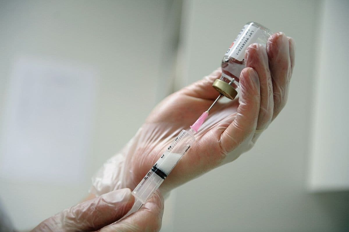 Mãos com luvas descartáveis puxam líquido de um frasco de vidro com uma seringa de agulha rosa